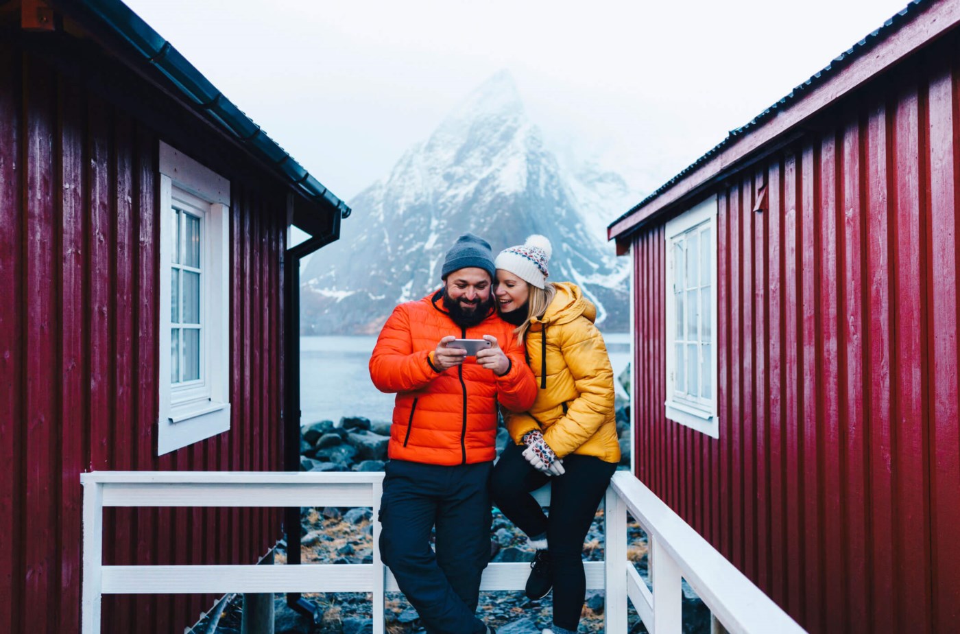 Et par i vinterklær ler sammen mens de ser på en mobiltelefon, stående mellom tradisjonelle røde bygg med en majestetisk, snødekt fjelltopp i bakgrunnen.