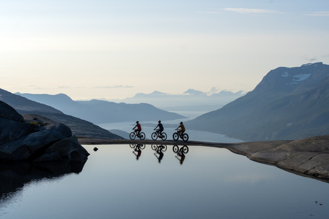 Tre syklister i Skjomen med fjell i bekgrunn og speilblankt vann
